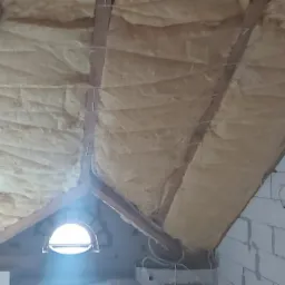 Poddasze w trakcie ocieplania, widoczna wełna mineralna między krokwiami, małe okrągłe okno w ścianie szczytowej, przewody elektryczne na ścianie z bloczków.