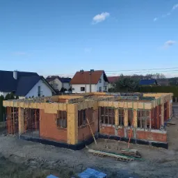 Stan surowy otwarty domu z czerwonej cegły w Wilkowie, widoczne szalunki i podpory, w tle zabudowa jednorodzinna i błękitne niebo.