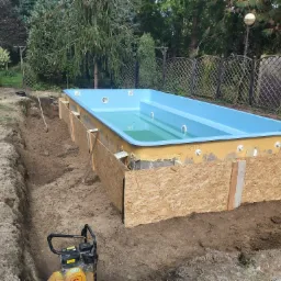 Montaż basenu w ogrodzie: wykopany dół, częściowo oszalowany, wypełniony wodą, widoczna zagęszczarka do gruntu.