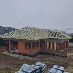 Budowa parterowego domu murowanego w stanie surowym otwartym z drewnianą konstrukcją dachu, widoczne bloczki ceramiczne i podpory szalunkowe, na pierwszym planie palety z materiałami budowlanymi.