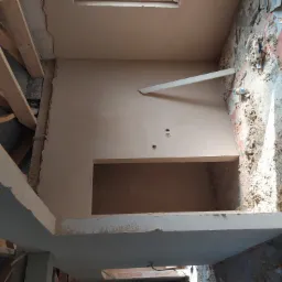 Wnętrze w trakcie remontu: ściana po nałożeniu gładzi gipsowej, widoczne instalacje elektryczne, fragmenty surowej ściany z resztkami zaprawy oraz drewniana konstrukcja stropu.