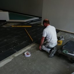 Lejkbud - Mężczyzna w białej koszulce i spodniach roboczych klęczy układając ciemne, prostokątne płytki na podłodze w pomieszczeniu z basenem; widoczne poziomice, puszka z narzędziami i wózek z zapasem płytek.