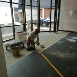 Lejkbud - Mężczyzna na kolanach z kielnią nakłada klej na podłogę pod płytki w lokalu z dużymi oknami, widoczne płytki, poziomica, wiadro z klejem i wózkiem z płytkami.