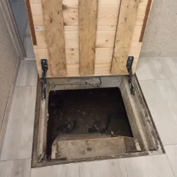 Otwarty właz do piwnicy z drewnianą klapą wykonaną z desek, widoczne schody w dół, jasne panele podłogowe dookoła.