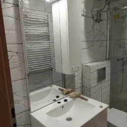 Biała umywalka z szafką, baterią i lustrem w łazience wykończonej płytkami imitującymi marmur, na umywalce leży gumowy młotek.