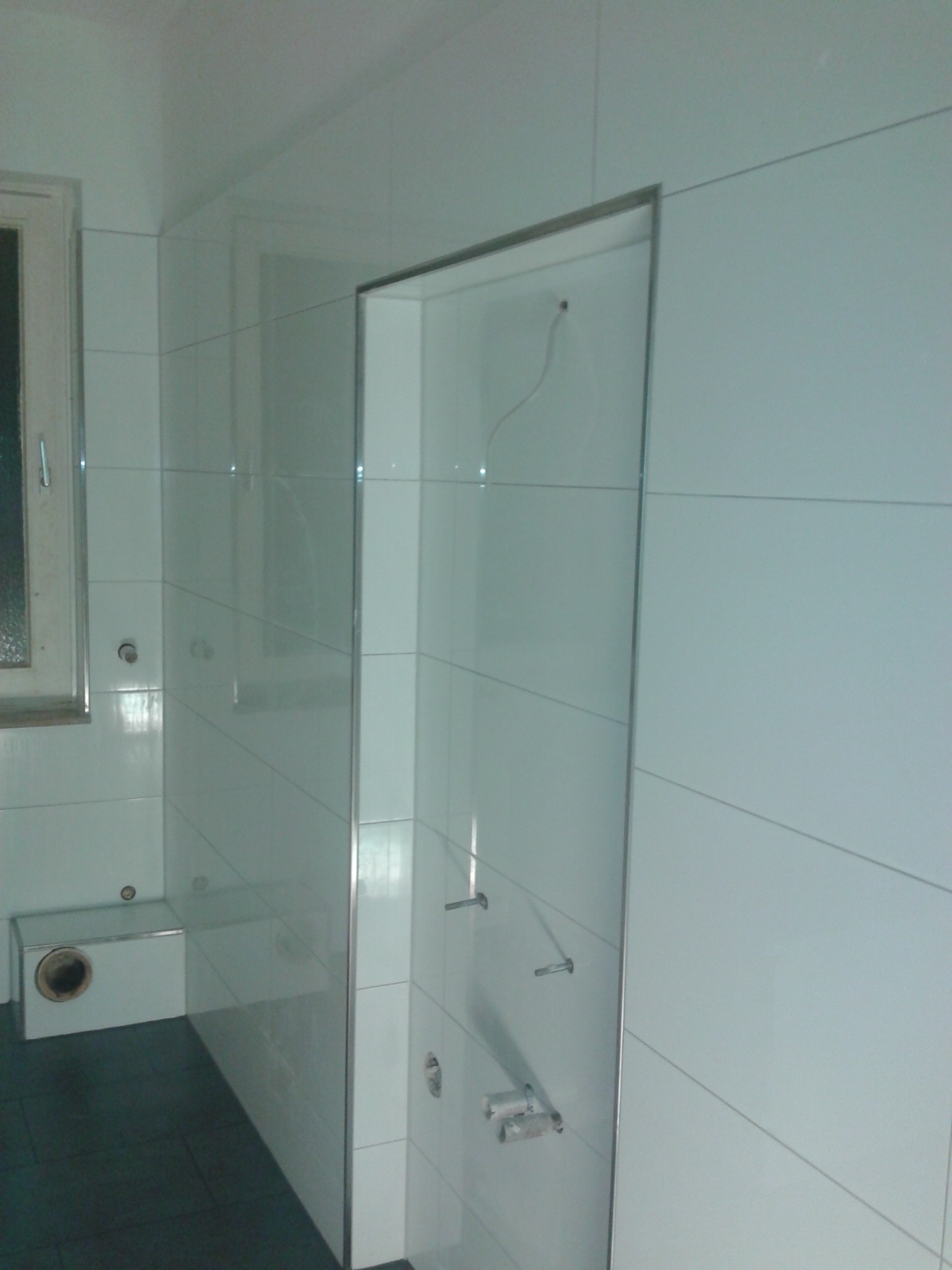 Świeżo wykafelkowana łazienka z widocznymi punktami mocowania armatury i niedokończonymi fugami, ciemna podłoga i okno z widocznym fragmentem ramy.