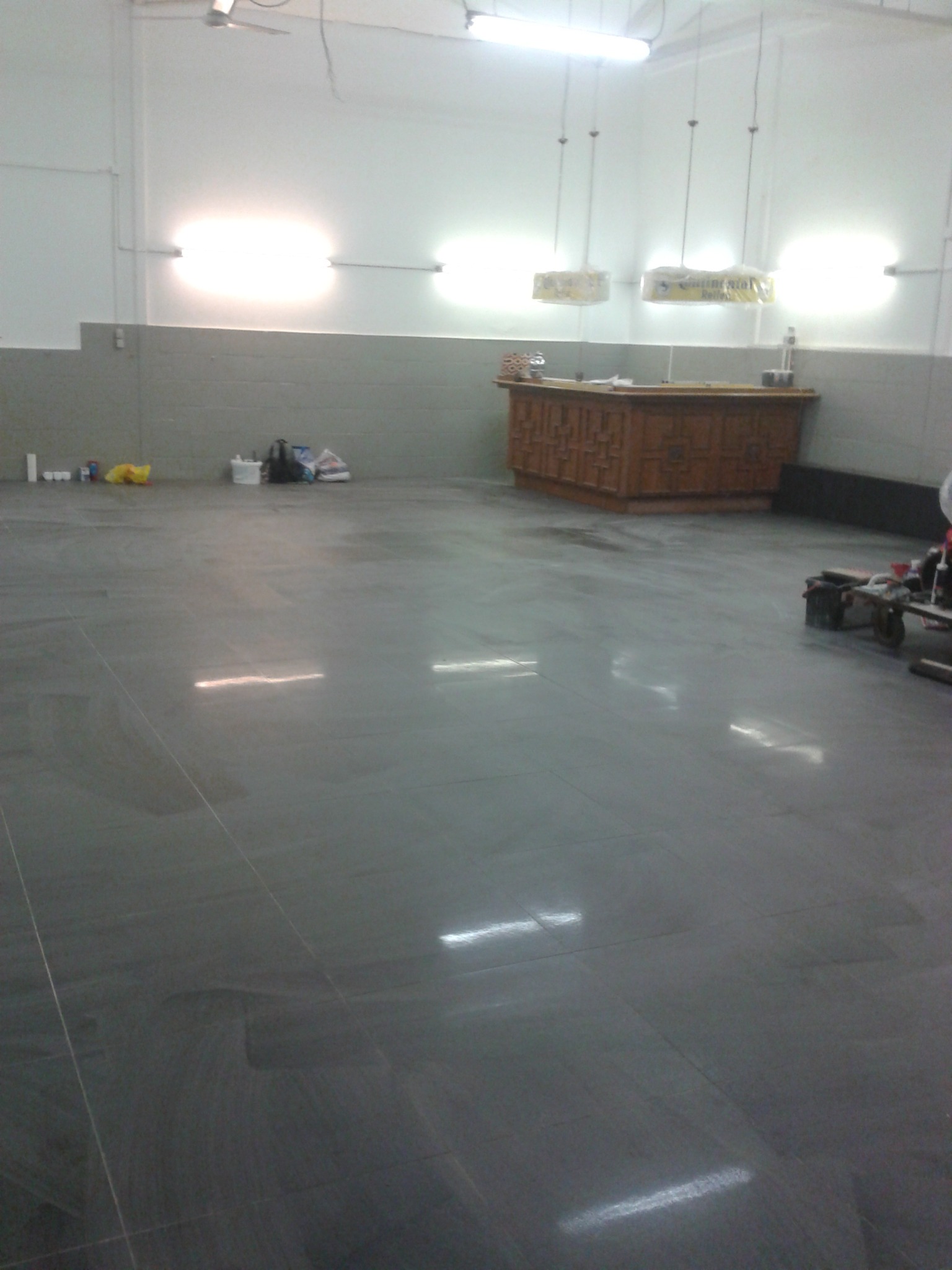 Wyłożona ciemnoszarą płytką podłoga w dużym, słabo oświetlonym pomieszczeniu, widoczne narzędzia i materiały budowlane w tle.