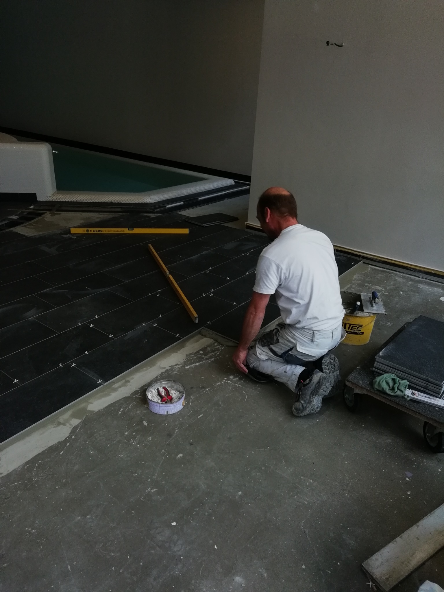 Mężczyzna w białej koszulce i spodniach roboczych klęczy układając ciemne, prostokątne płytki na podłodze w pomieszczeniu z basenem; widoczne poziomice, puszka z narzędziami i wózek z zapasem płytek.