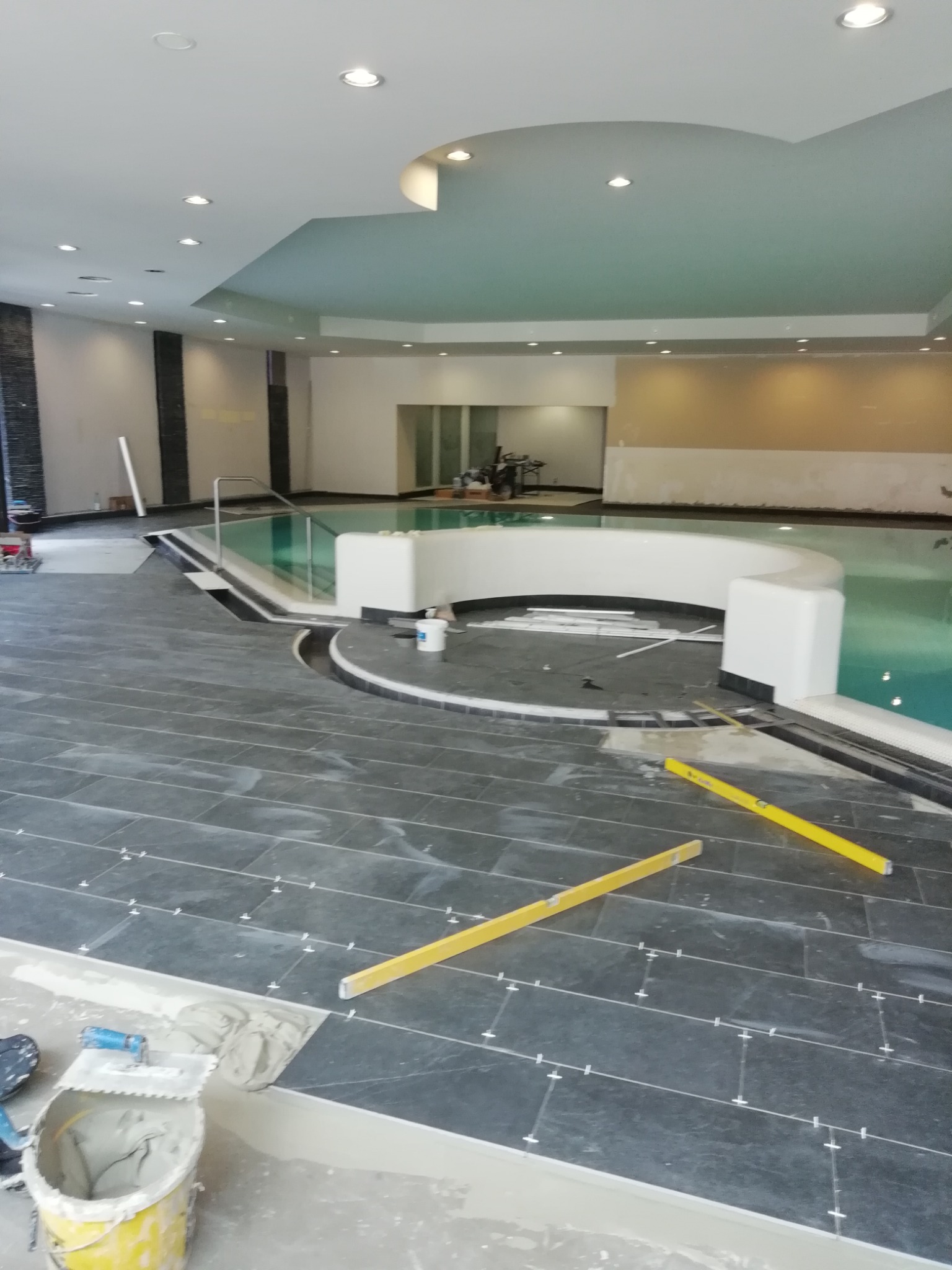 Układanie ciemnoszarej, prostokątnej glazury wzdłuż basenu, widoczne poziomice, krzyżyki dystansowe i wiadro z zaprawą.