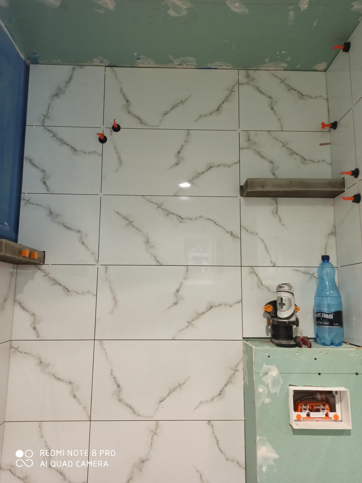 Białe płytki imitujące marmur na ścianie w trakcie układania, z systemem poziomowania płytek, widoczne narzędzia i butelka wody na tle surowej ściany z płyt gipsowo-kartonowych.
