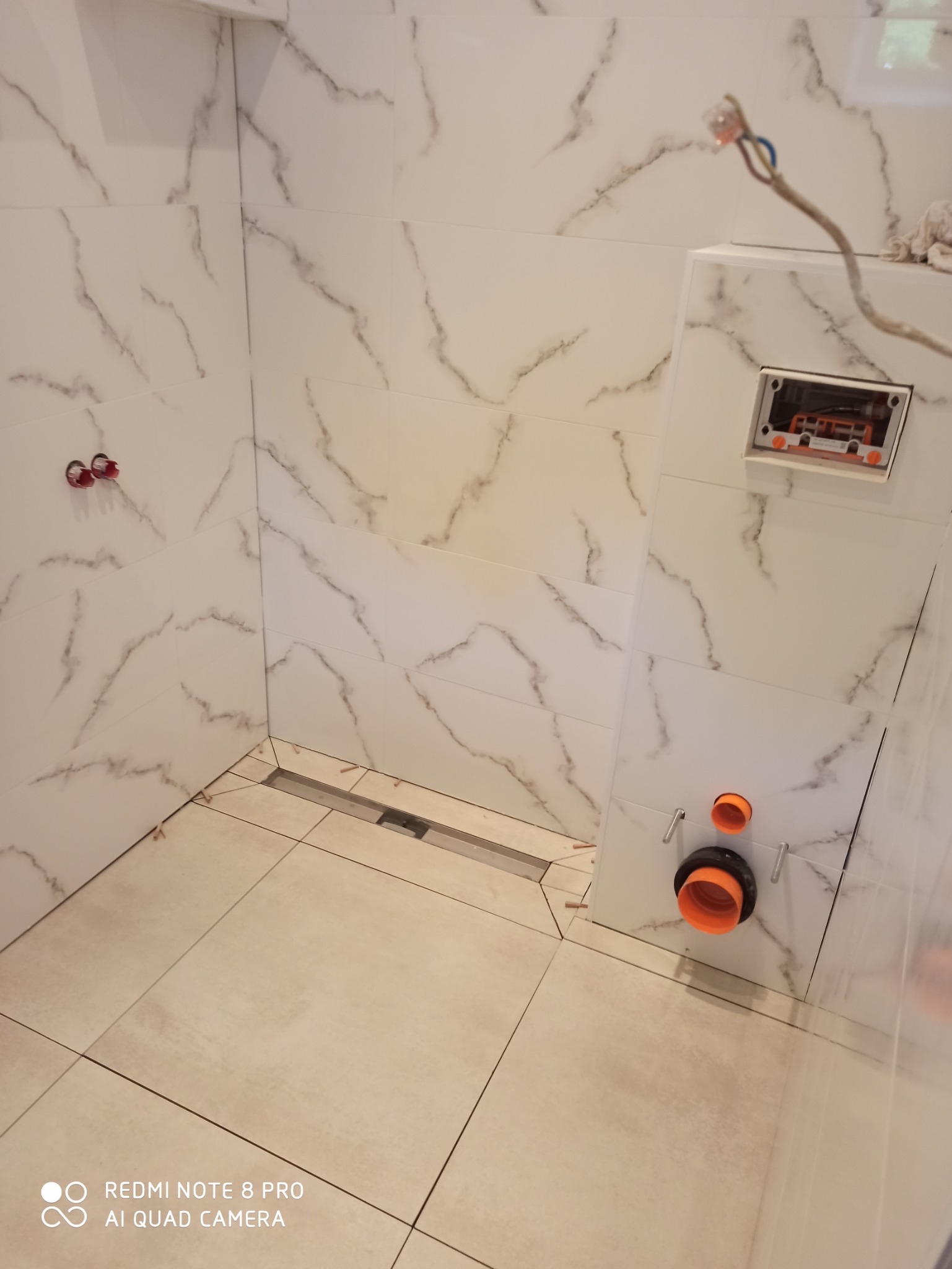 Wnętrze łazienki w trakcie remontu, widoczne białe płytki z marmurkowym wzorem na ścianach i podłodze, zamontowany odpływ liniowy oraz elementy instalacji podtynkowej.