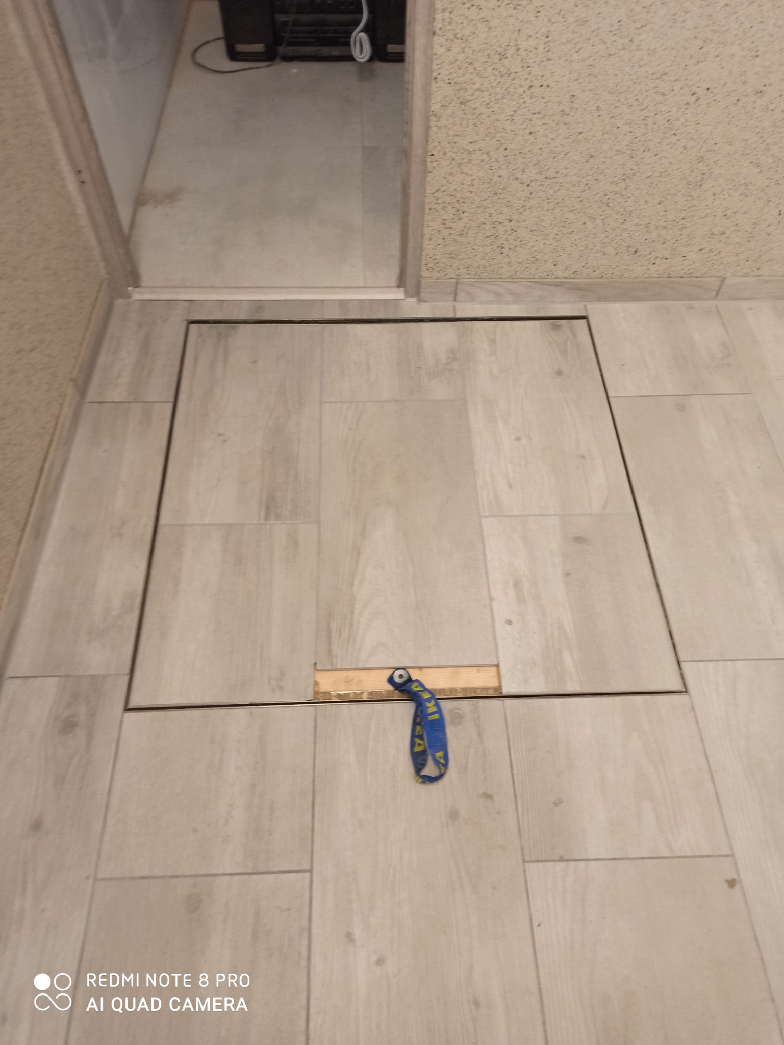 Szara podłoga wyłożona płytkami imitującymi drewno z ukrytym włazem rewizyjnym, otwartym przy pomocy drewnianego klocka i niebieskiej zawieszki IKEA.