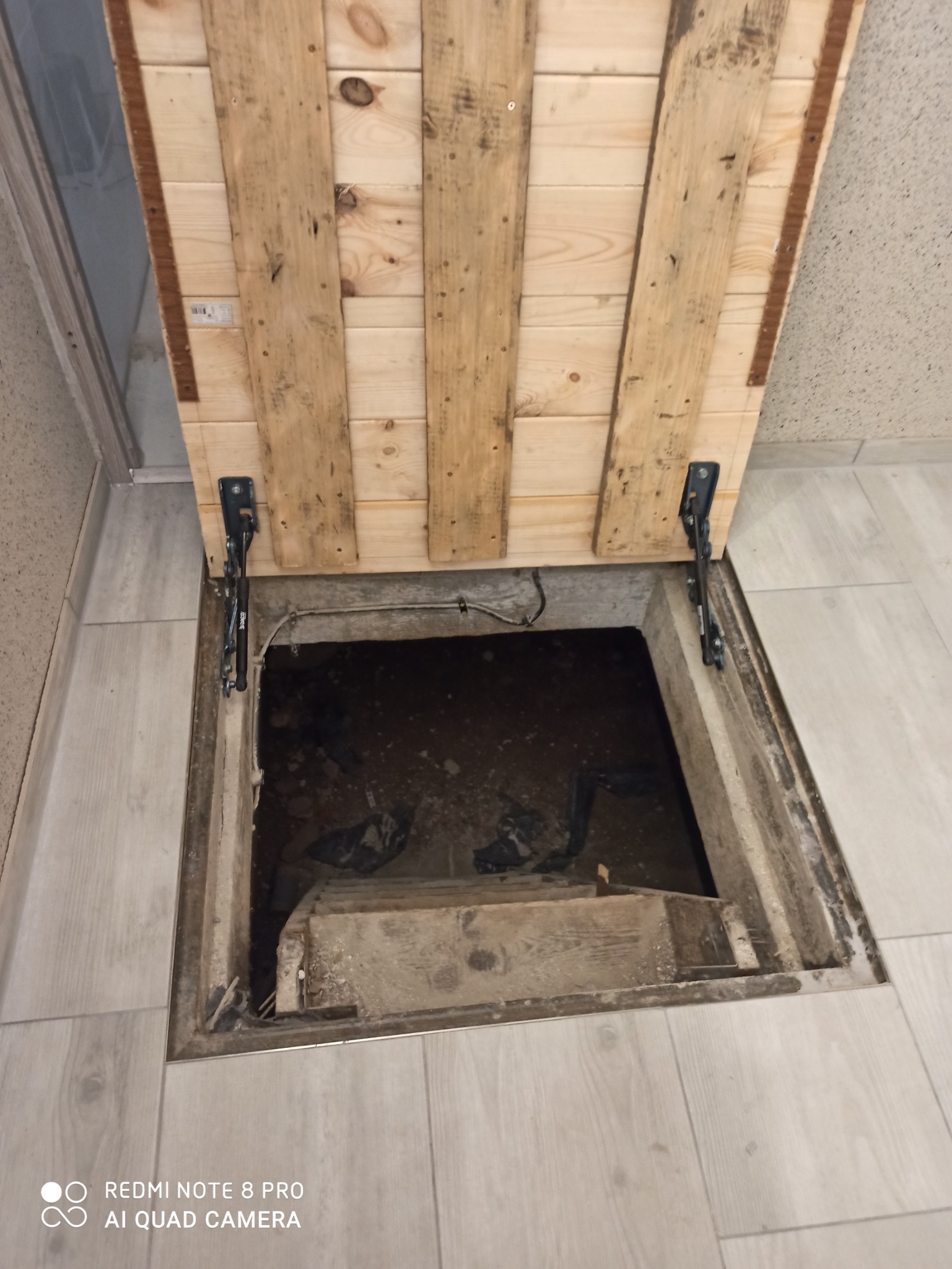 Otwarty właz do piwnicy z drewnianą klapą wykonaną z desek, widoczne schody w dół, jasne panele podłogowe dookoła.