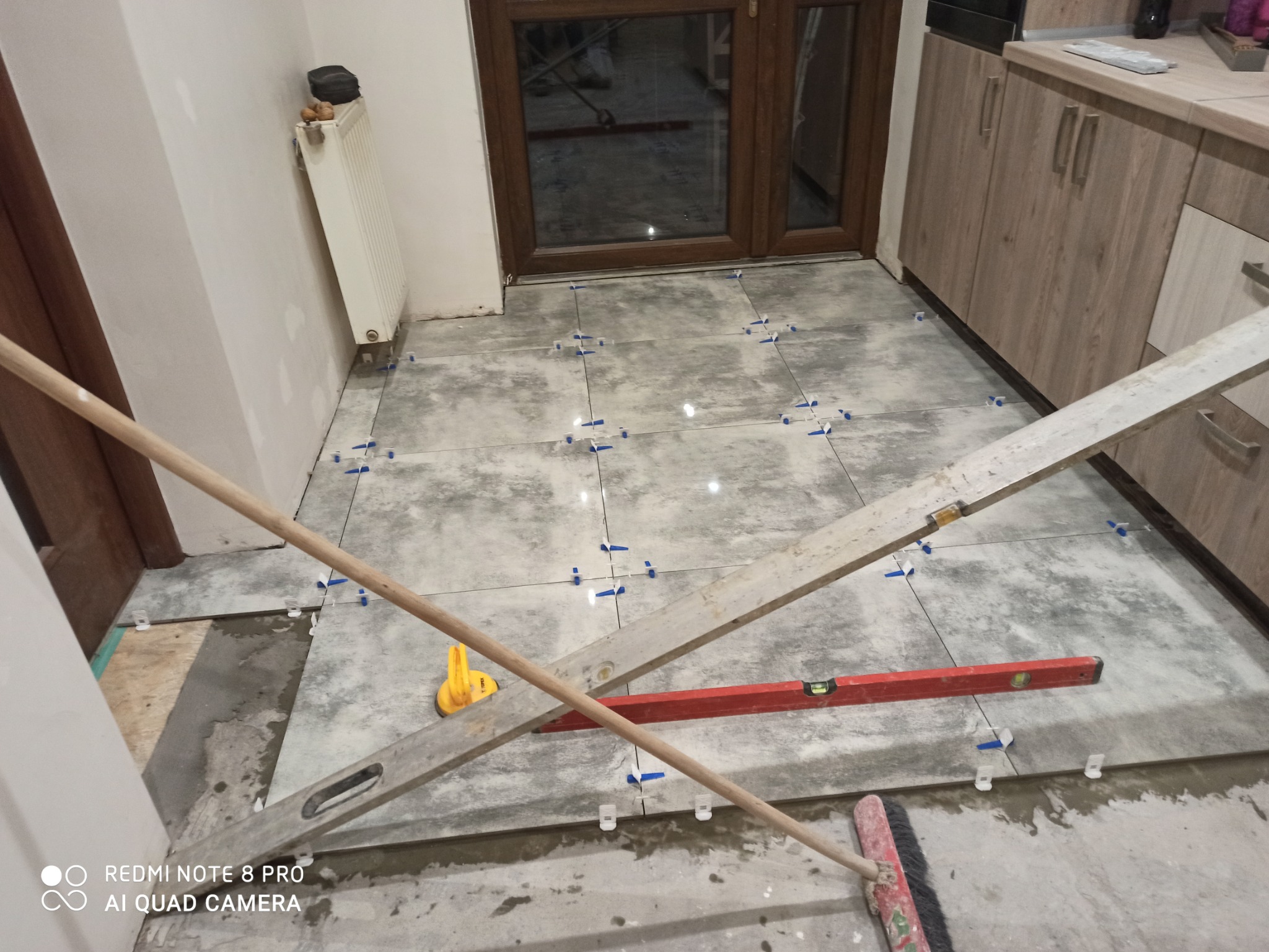 Układanie szarych płytek podłogowych w kuchni, widoczne krzyżyki dystansowe, poziomica i narzędzia glazurnicze, częściowo ułożona podłoga, widok na okno i zabudowę kuchenną