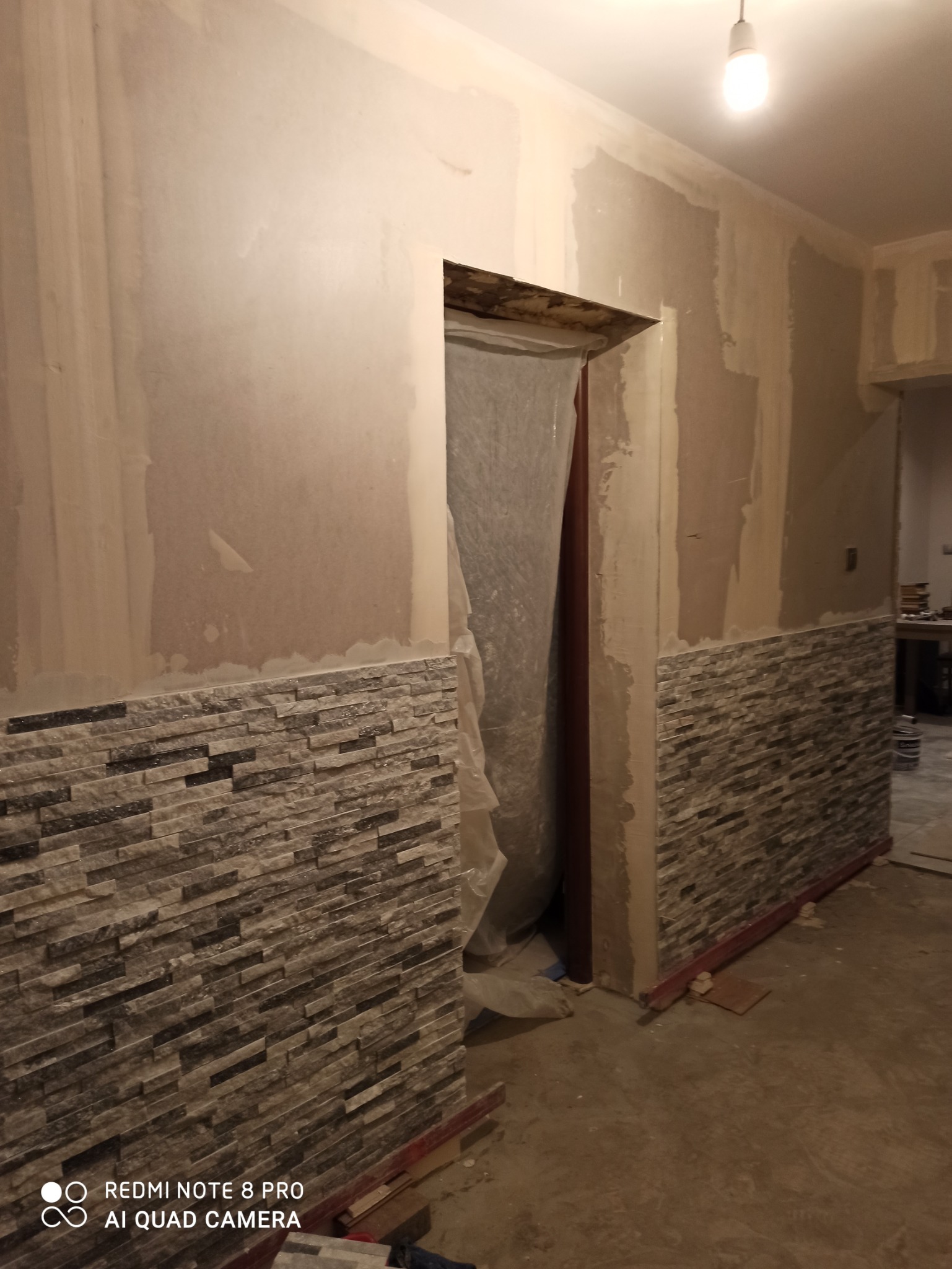 Wnętrze w trakcie remontu: ściany częściowo pokryte gładzią, częściowo wykończone dekoracyjną okładziną kamienną, zabezpieczone folią drzwi w przejściu.