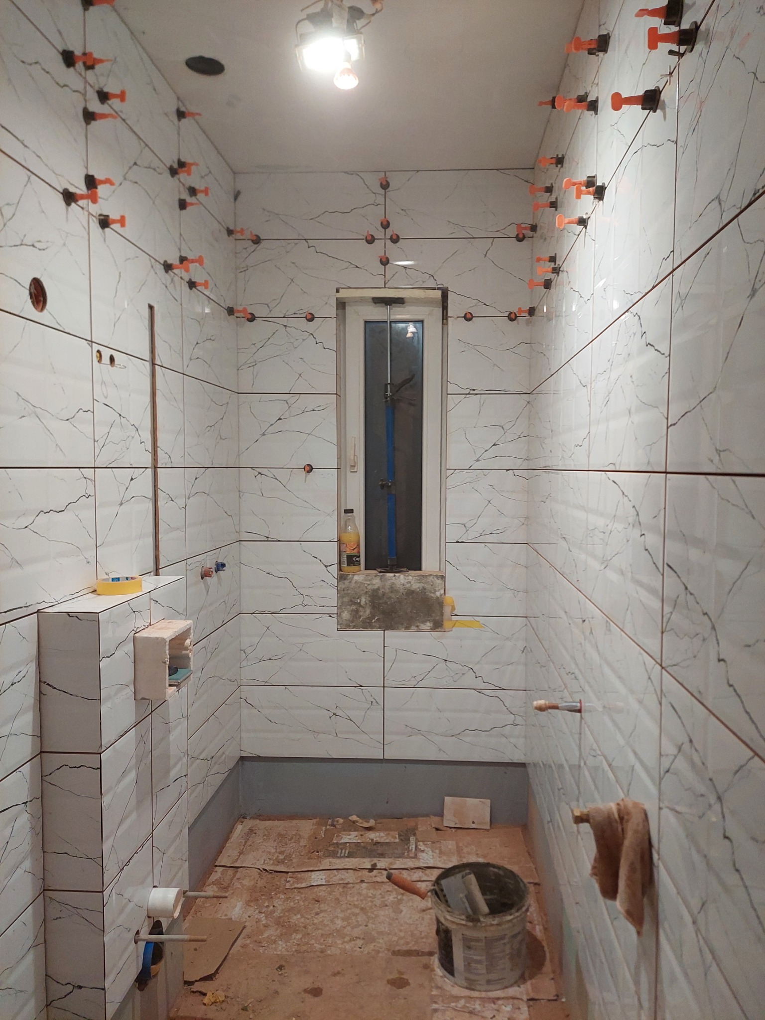 Łazienka w trakcie układania płytek imitujących marmur, z widocznymi systemami poziomowania płytek w kolorze pomarańczowym, narzędziami w wiadrze i fragmentem nieukończonej podłogi.
