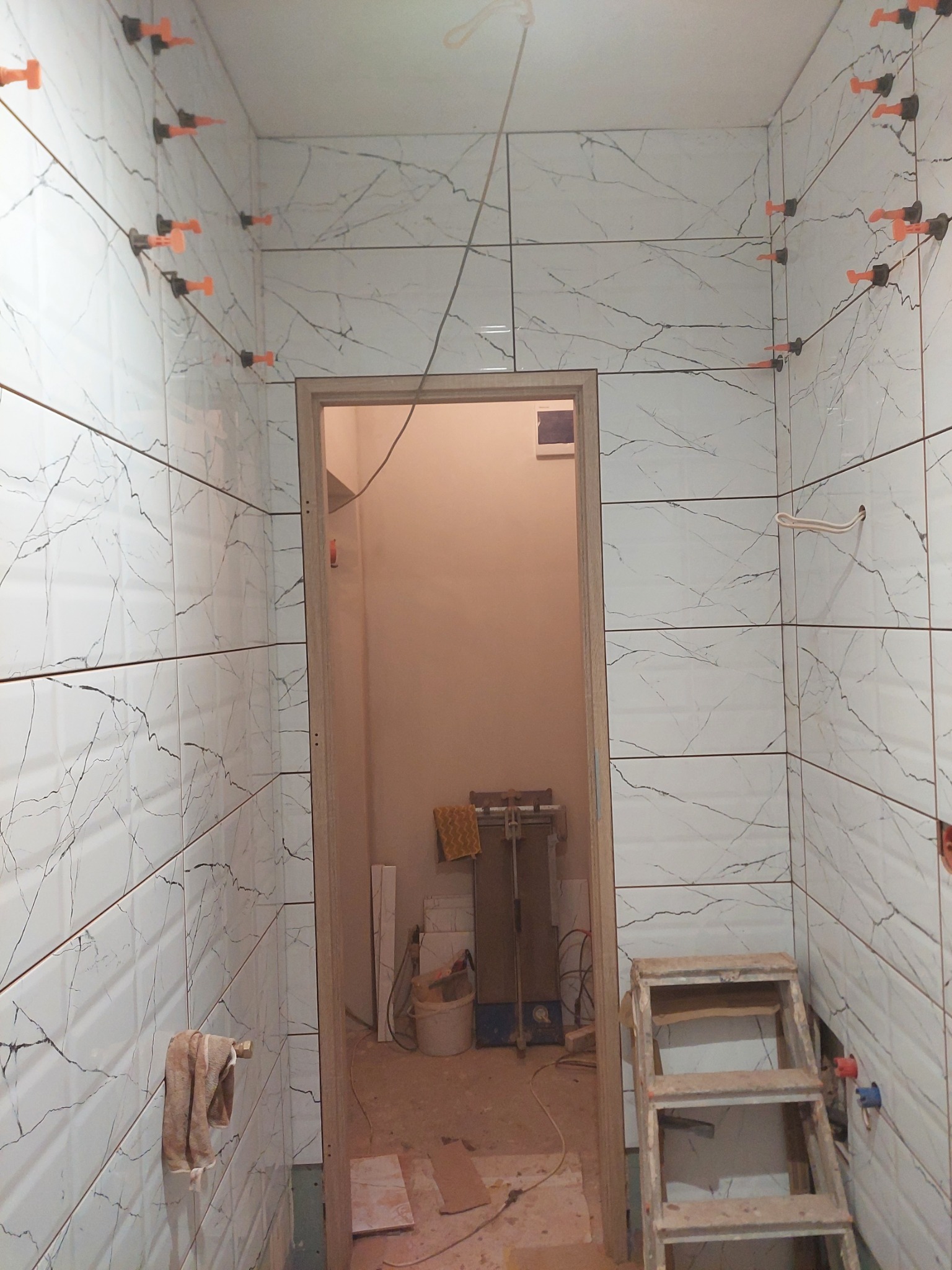 Łazienka w trakcie wykańczania: białe płytki imitujące marmur z ciemnymi żyłami, pomarańczowe klipsy poziomujące płytki, drabina, widok na sąsiednie pomieszczenie.