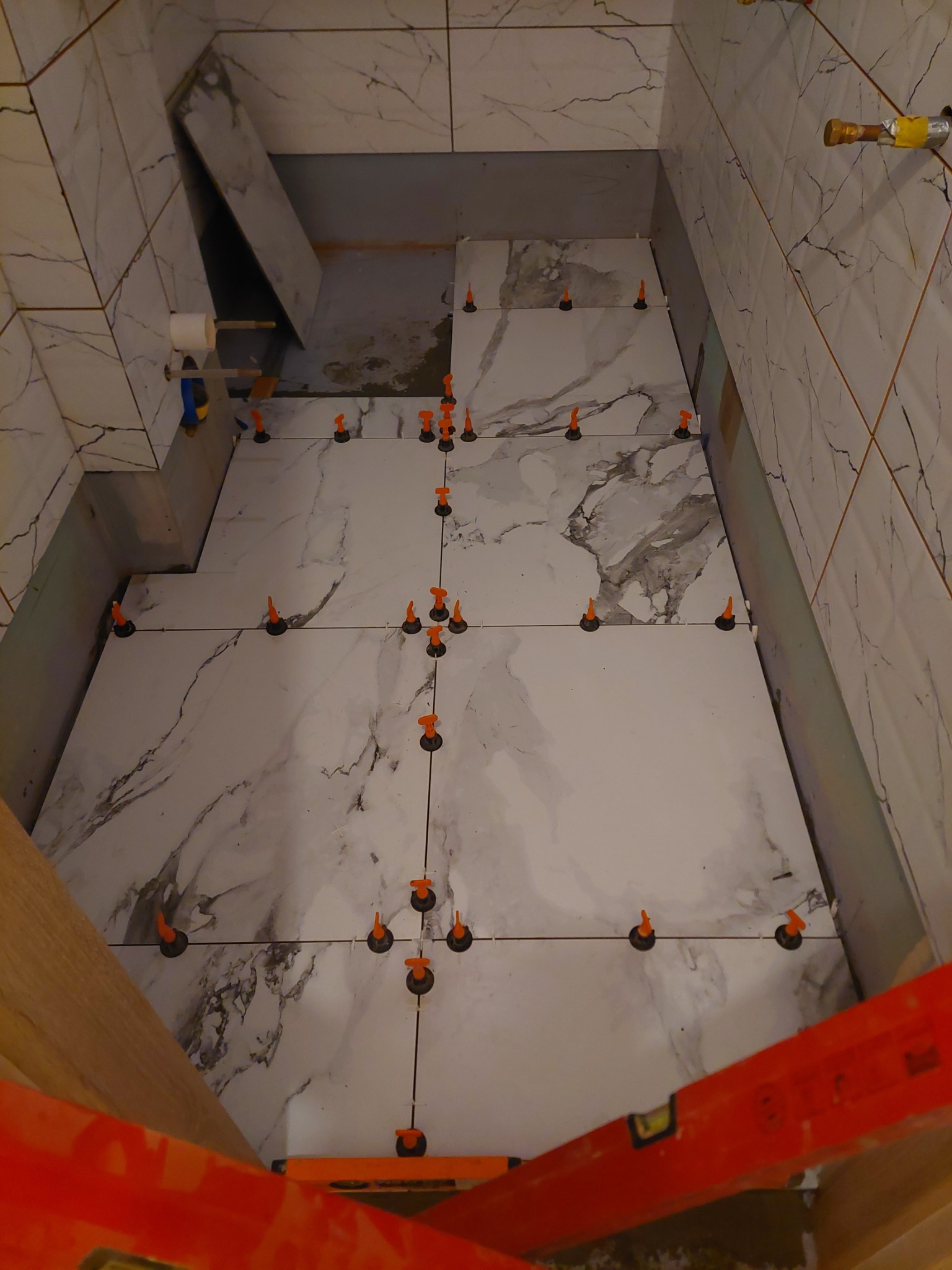 Układanie wielkoformatowych płytek imitujących marmur w łazience w Świerczynie 22/2, widoczne pomarańczowe klipsy poziomujące i fragment poziomicy, etap prac wykończeniowych.