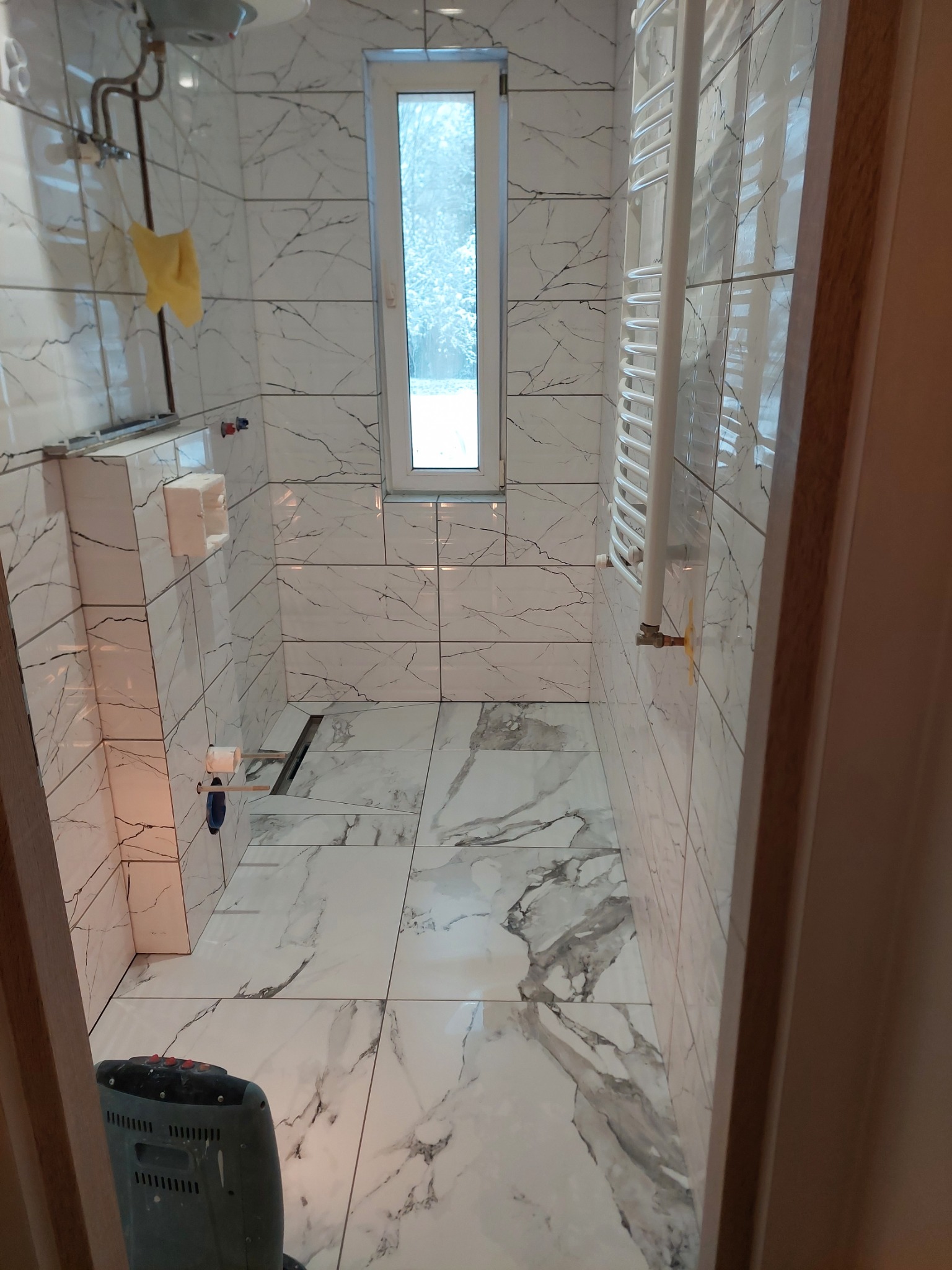 Wąska łazienka wykończona białymi płytkami imitującymi marmur, z grzejnikiem drabinkowym i wąskim oknem. Widoczny fragment grzejnika elektrycznego na podłodze.