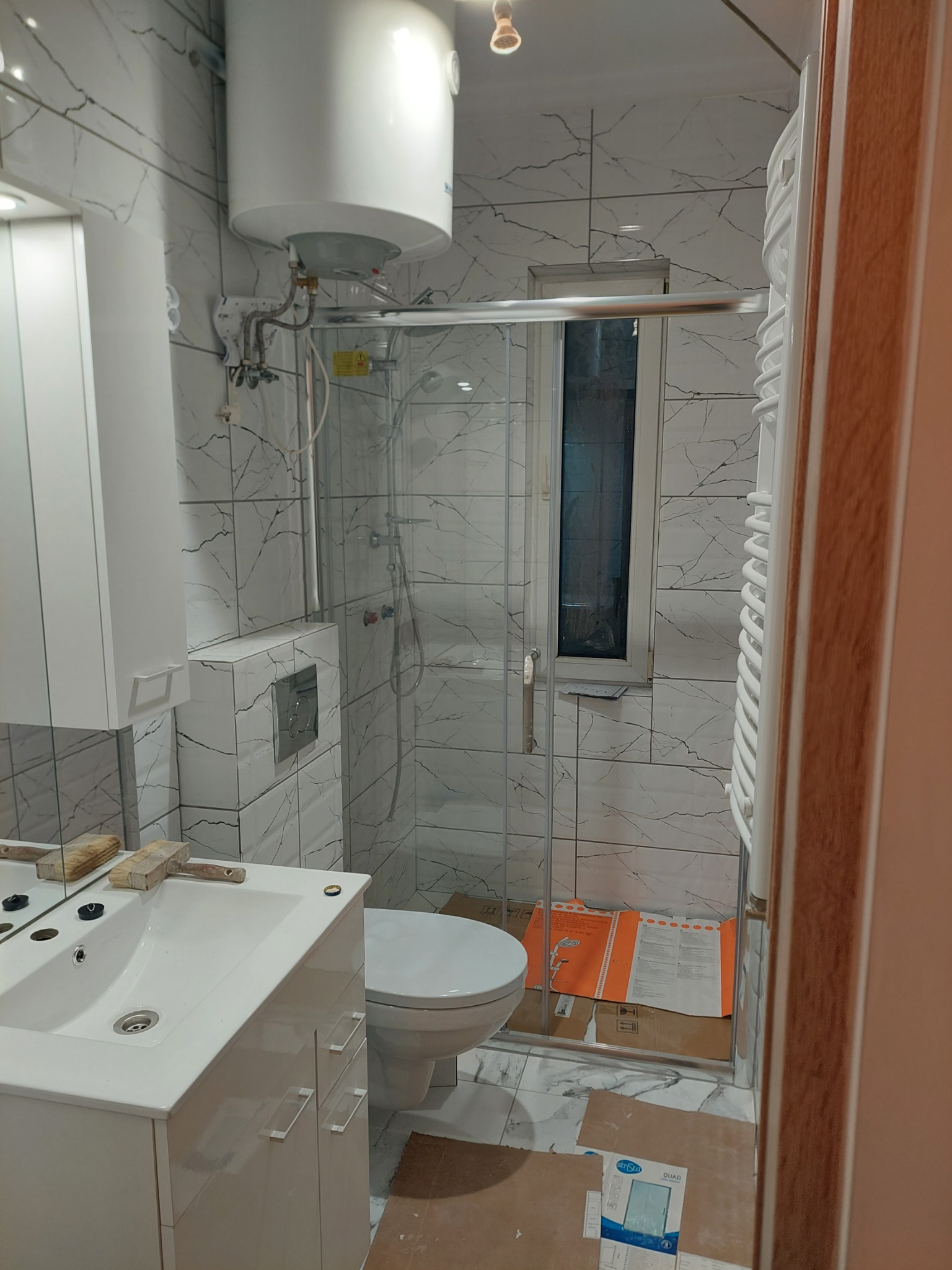 Remont łazienki w trakcie: białe płytki imitujące marmur, kabina prysznicowa ze szkła, biała umywalka z szafką i bojler elektryczny nad kabiną, widoczne kartony po płytkach.