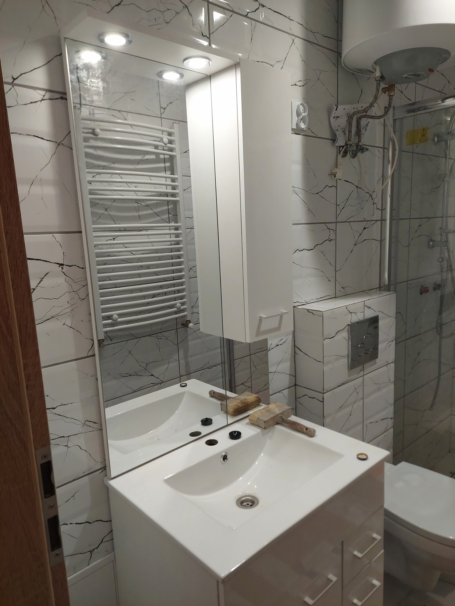 Biała umywalka z szafką, baterią i lustrem w łazience wykończonej płytkami imitującymi marmur, na umywalce leży gumowy młotek.