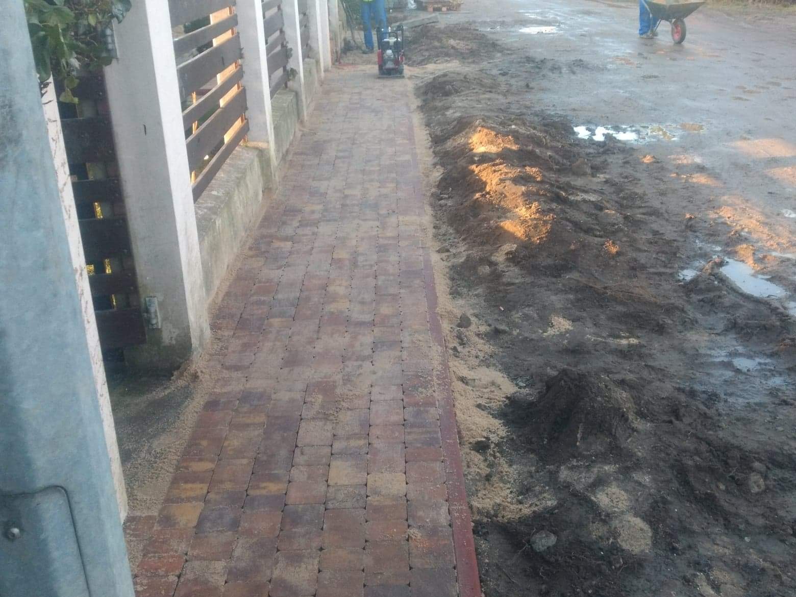 Układanie kostki brukowej: zagęszczarka wibrująca ubija nowo ułożoną, brązowo-czerwoną kostkę brukową, obok przygotowany teren z błotem i w oddali pracownik z taczką.