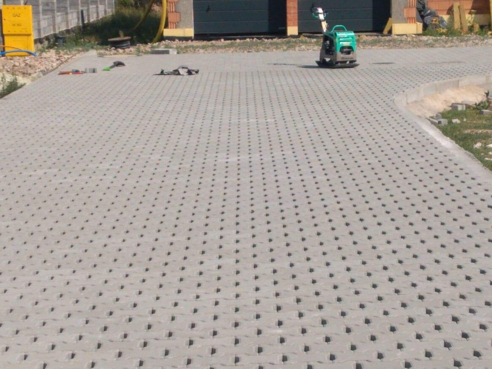 Ułożona szara ażurowa nawierzchnia z betonu z widoczną ubijarką wibracyjną i narzędziami brukarskimi w tle.