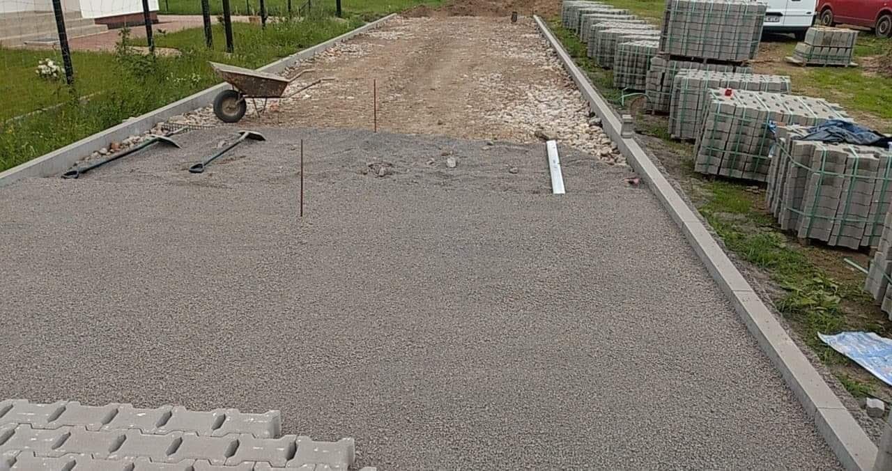 Przygotowanie podłoża pod nawierzchnię: wyrównany żwir, obrzeża betonowe, stosy kostki, taczka i narzędzia w tle.