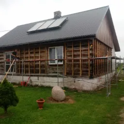 Dom z bali w trakcie prac elewacyjnych: widoczne rusztowania, częściowo odsłonięta konstrukcja drewniana, przygotowania do ocieplenia, panele solarne na dachu.