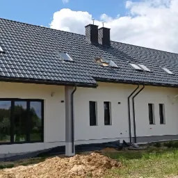 Nowoczesny dom jednorodzinny z ciemną dachówką, jasną elewacją i czarnymi oknami, widok z zewnątrz w słoneczny dzień.