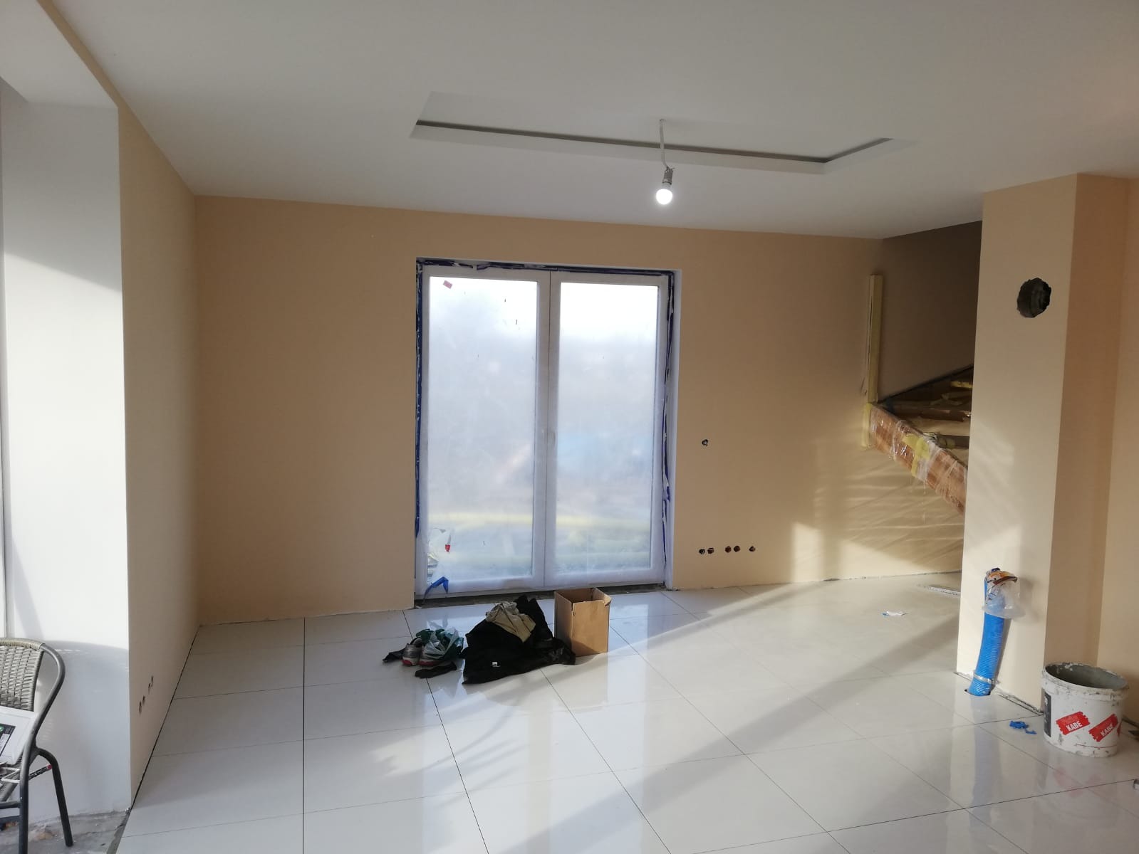 Wnętrze w trakcie prac wykończeniowych: świeżo położone białe płytki podłogowe, ściany w odcieniach beżu, okno zabezpieczone folią malarską, widoczne narzędzia i materiały budowlane, przygotowane...