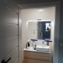 Nowoczesna łazienka z umywalką nablatową na drewnianej szafce, widok zza uchylonych drzwi, z odbiciem osoby robiącej zdjęcie w lustrze.