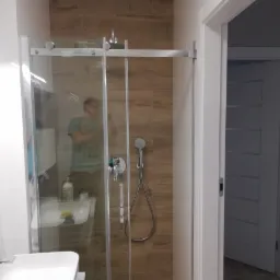 Szklana kabina prysznicowa z przesuwnymi drzwiami, drewnopodobne płytki na ścianie wewnątrz kabiny, widoczna umywalka z niebieską gąbką, białe płytki na pozostałej części ściany.