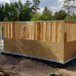 Wznoszenie drewnianej konstrukcji szkieletowej domu na fundamencie, widoczne płyty OSB na dolnej części ścian, teren piaszczysty z elementami zieleni.