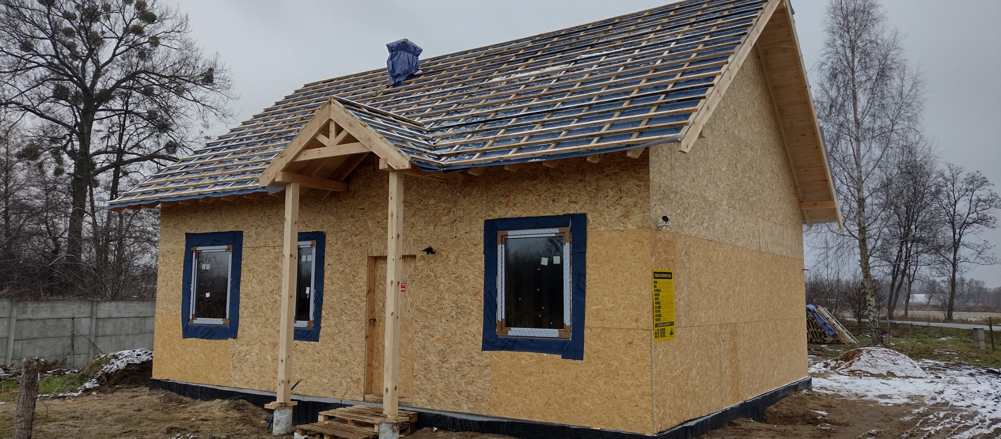 Surowy stan domu z drewnianych płyt OSB z widoczną konstrukcją dachu i okien z niebieską folią zabezpieczającą, na działce z resztkami śniegu i betonowym ogrodzeniem.