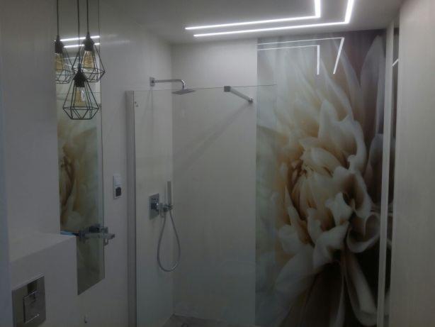 Nowoczesna łazienka z przeszkloną kabiną prysznicową ozdobioną grafiką kwiatową, geometryczne lampy wiszące i oświetlenie LED na suficie.
