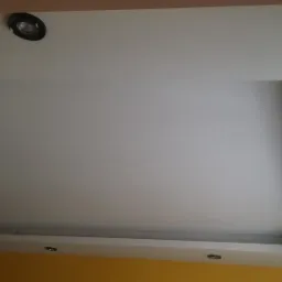 Biały sufit podwieszany z wbudowanymi reflektorami, widok z dołu, fragment żółtej ściany w tle.