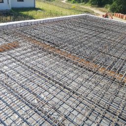 Skibicki Adrian - Zbrojenie płyty fundamentowej z widocznymi prętami i siatką stalową, przygotowane do zalania betonem, na tle budynku w budowie i zielonego terenu.