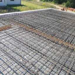 Zbrojenie płyty fundamentowej z widocznymi prętami i siatką stalową, przygotowane do zalania betonem, na tle budynku w budowie i zielonego terenu.