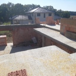 Skibicki Adrian - Wznoszenie ścian parteru z pustaków ceramicznych na budowie domu jednorodzinnego, widoczne zbrojenia nadproży, w tle drugi budynek w stanie surowym otwartym.