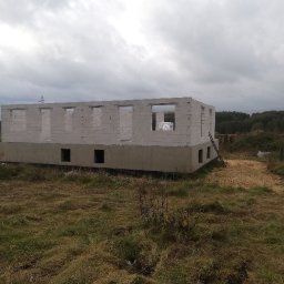 Skibicki Adrian - Surowy stan domu z betonu komórkowego na fundamencie, widoczne otwory okienne i drzwiowe, drabina oparta o ścianę, w tle zalesiony teren i pochmurne niebo.