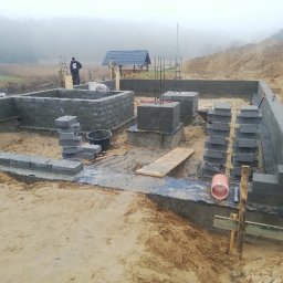 Skibicki Adrian - Budowa fundamentów domu z bloczków betonowych, widoczne zbrojenia, teren budowy z piaskiem i ziemią, w tle zalesiony teren i mgła.