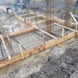 Skibicki Adrian - Wylane fundamenty pod budowę domu, widoczne drewniane szalunki, zbrojenie z prętów stalowych i świeży beton, kilku pracowników w tle.