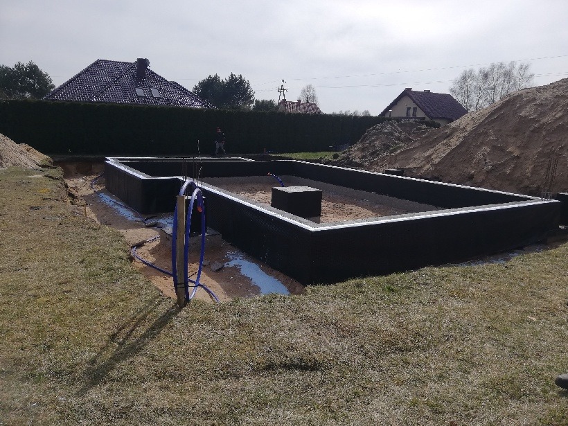 Zabezpieczony fundament pod budynek mieszkalny, widoczne izolacje pionowe, teren wokół wyrównany, na dalszym planie dom jednorodzinny i kopiec ziemi.