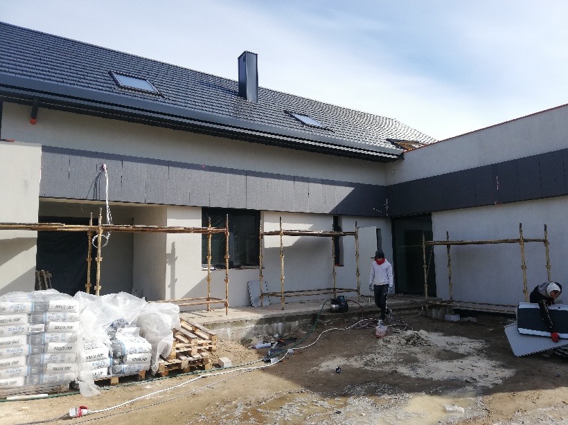 Nowoczesny dom w trakcie budowy, z szarymi panelami elewacyjnymi, widoczne rusztowania, worki z zaprawą i pracownicy na placu budowy.