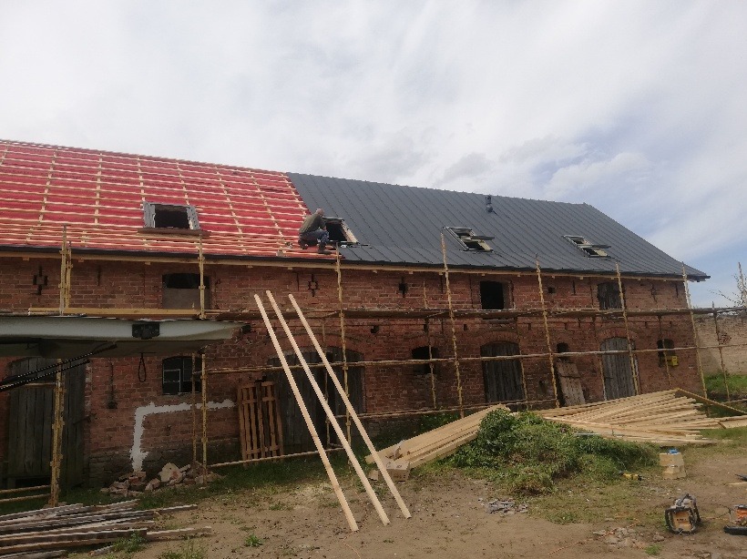 Renowacja dachu dwukondygnacyjnego budynku z czerwonej cegły, część dachu pokryta czerwoną dachówką, część czarną blachą, widoczne rusztowania i pracownik przy oknie dachowym.