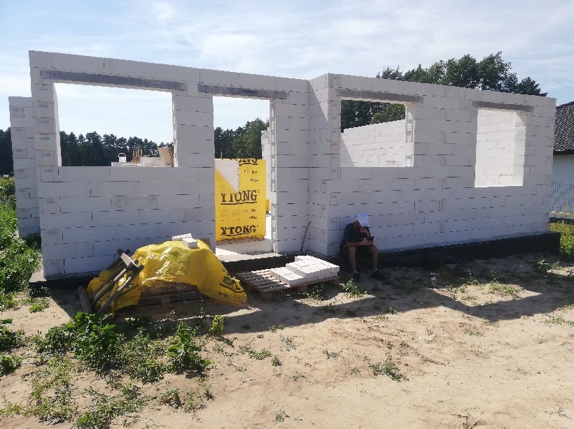 Wznoszona ściana domu z białych bloczków Ytong, z widocznymi otworami okiennymi, paletą bloczków i folią ochronną, na zewnątrz siedzi osoba z telefonem.