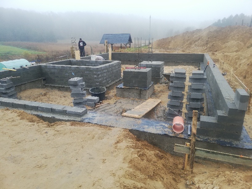 Budowa fundamentów domu z bloczków betonowych, widoczne zbrojenia, teren budowy z piaskiem i ziemią, w tle zalesiony teren i mgła.