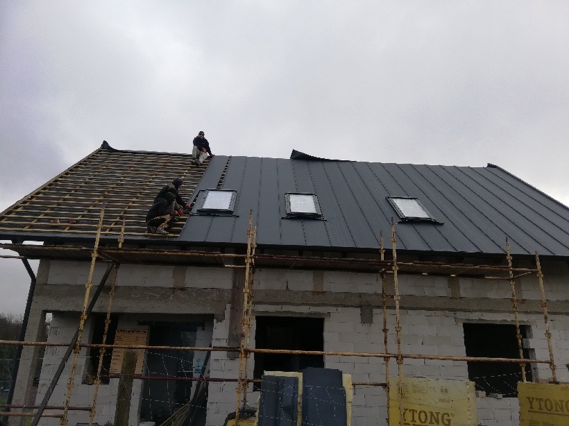 Montaż blachy na dachu domu w stanie surowym otwartym, widoczne okna dachowe i rusztowanie.