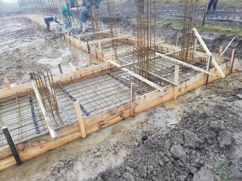 Wylane fundamenty pod budowę domu, widoczne drewniane szalunki, zbrojenie z prętów stalowych i świeży beton, kilku pracowników w tle.